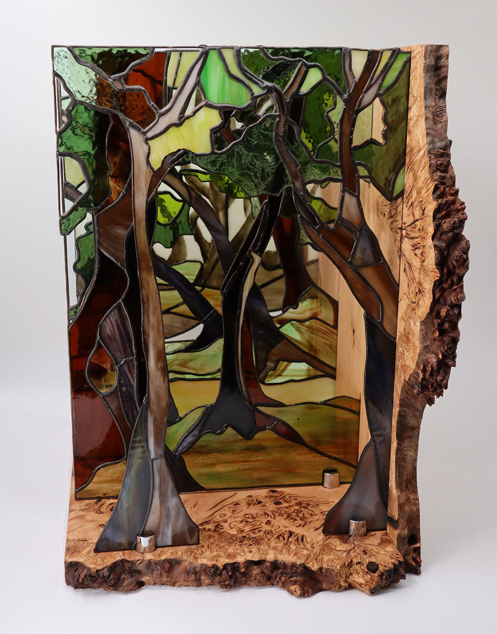 A stained glass art piece depicts a forest scene with tall trees, framed by a textured wooden border that resembles natural bark. The glass is in various shades of green and brown, creating a vibrant and earthy image.