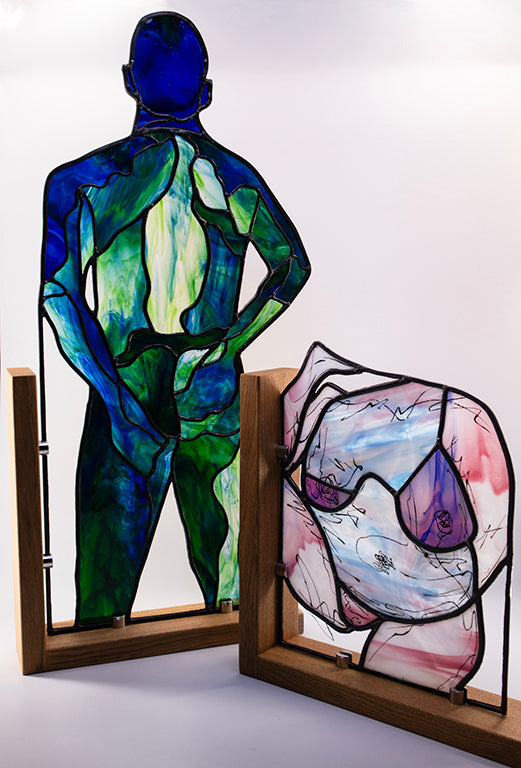 Stained glass artwork depicting a standing figure in blue and green tones and a reclining abstract figure in pink and white, set in wooden frames on a white background.