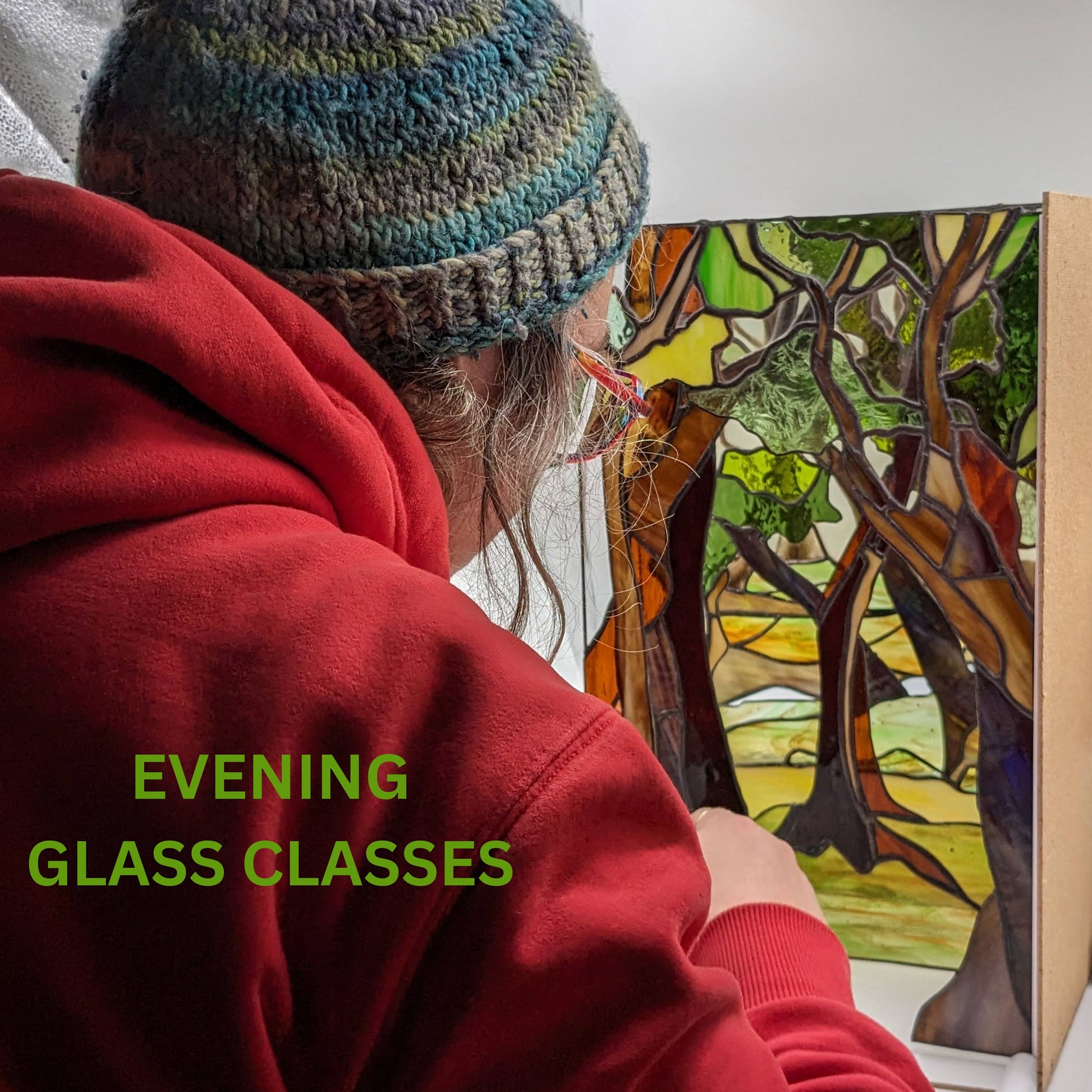 A person in a red hoodie and knit hat meticulously crafts a stained glass piece depicting trees, demonstrating expert leaded light techniques. Elegantly overlaid on the image are the words: "SPRING 10 Week Stained Glass Course (In Person), Tuesday EVENING by LF Kaleidoscope.