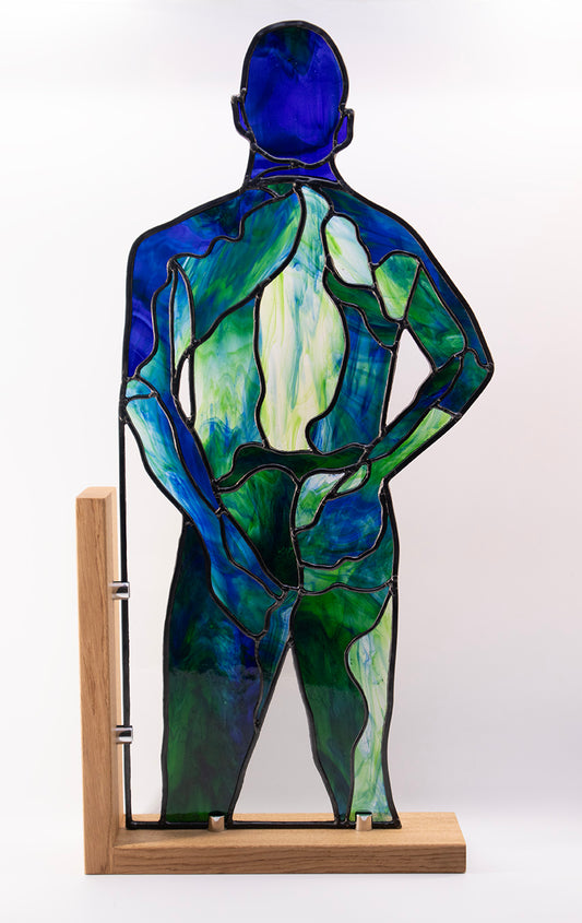 A luxurious stained glass artwork depicts a human figure from the back in serene shades of blue and green. The figure stands confidently on a wooden base, hands resting on the hips. This semi-transparent piece, perfect for collectors, allows light to pass through beautifully. An exceptional example of skilled craftsmanship- this artwork was cut from a single sheet of glass.