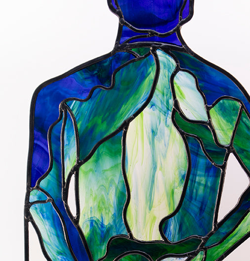 Close up of the shoulders and back: 
A luxurious stained glass artwork depicts a human figure from the back in serene shades of blue and green. The figure stands confidently on a wooden base, hands resting on the hips. This semi-transparent piece, perfect for collectors, allows light to pass through beautifully. An exceptional example of skilled craftsmanship- this artwork was cut from a single sheet of glass.