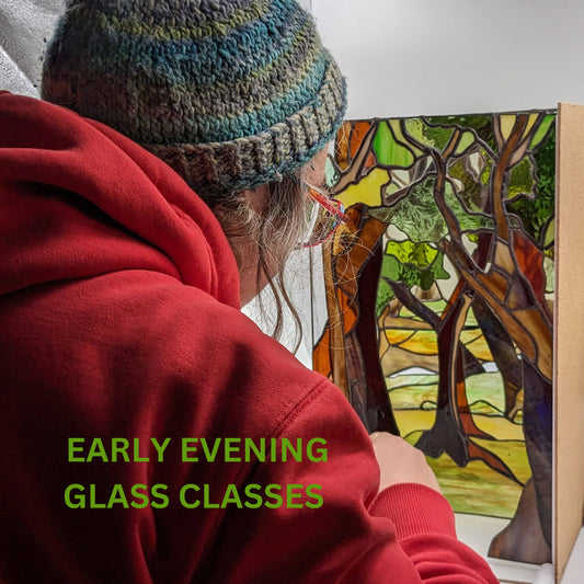 In a stained glass studio, an individual clad in a red hoodie and blue knit hat is focused on creating a vibrant stained glass panel featuring trees. The text reads, "10 Week Stained Glass Course (In Person), Tuesday EVENING by LF Kaleidoscope. Explore copper foiling, leaded stained glass and glass painting. 