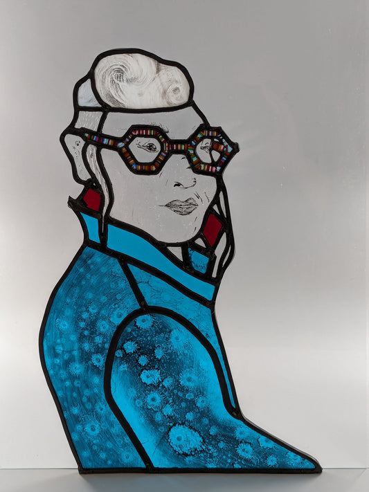 The product "Framed on the Train (Self Portrait)" by LF Kaleidoscope features a stained-glass artwork portraying an individual with short hair, fashionable large glasses, and vivid red earrings. They are dressed in a striking blue jacket embellished with detailed circular motifs. This self portrait is made entirely from glass and is a unique artifact.