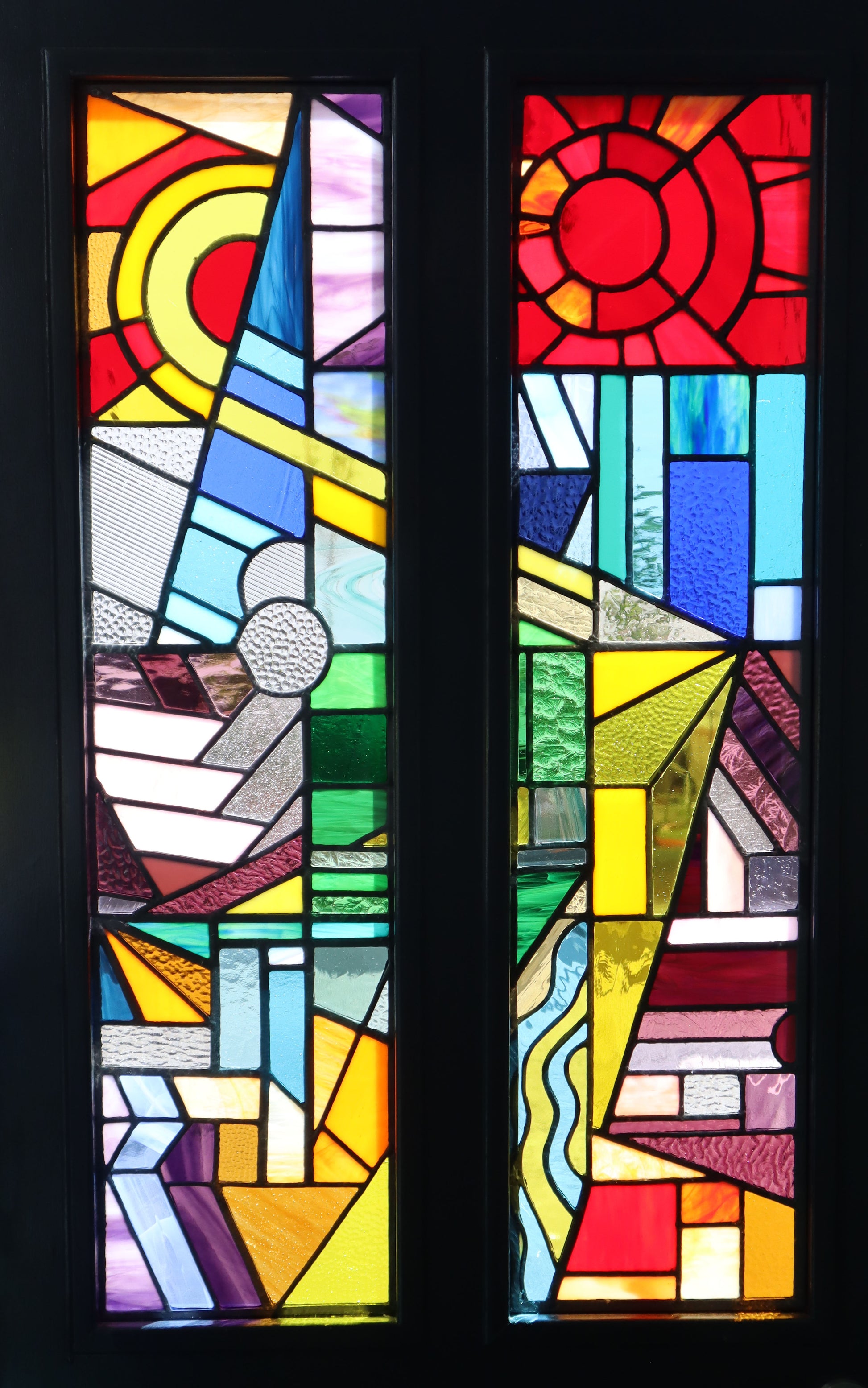 LF Kaleidoscope's Front Door and window commissions, combine cutting edge craftsmanship with leading encapsulating and safety standards to create bespoke stained glass replacement windows. The examples here are of vibrant geometric patterns. This design showcases circles, triangles, and rectangles in a spectrum of red, yellow, blue, green, purple, and white hues. The abstract shapes and lines come together to form a stunning colorful patchwork effect that creates a feature for any property. All bespoke. 