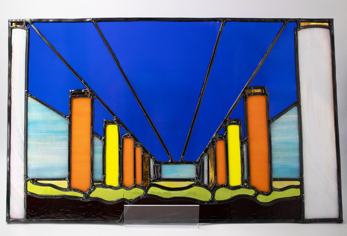 The artwork titled "Neon Fly Over" by LF Kaleidoscope features a stained glass design with geometric forms, including vertical orange and yellow columns set against a deep blue overpass. Black leadlines converge towards the horizon, evoking the perspective of a neon flyover, while the lower section showcases undulating green and purple glass. A playful contemporary artwork celebrating construction and industry artwork, that would look great in any office. 