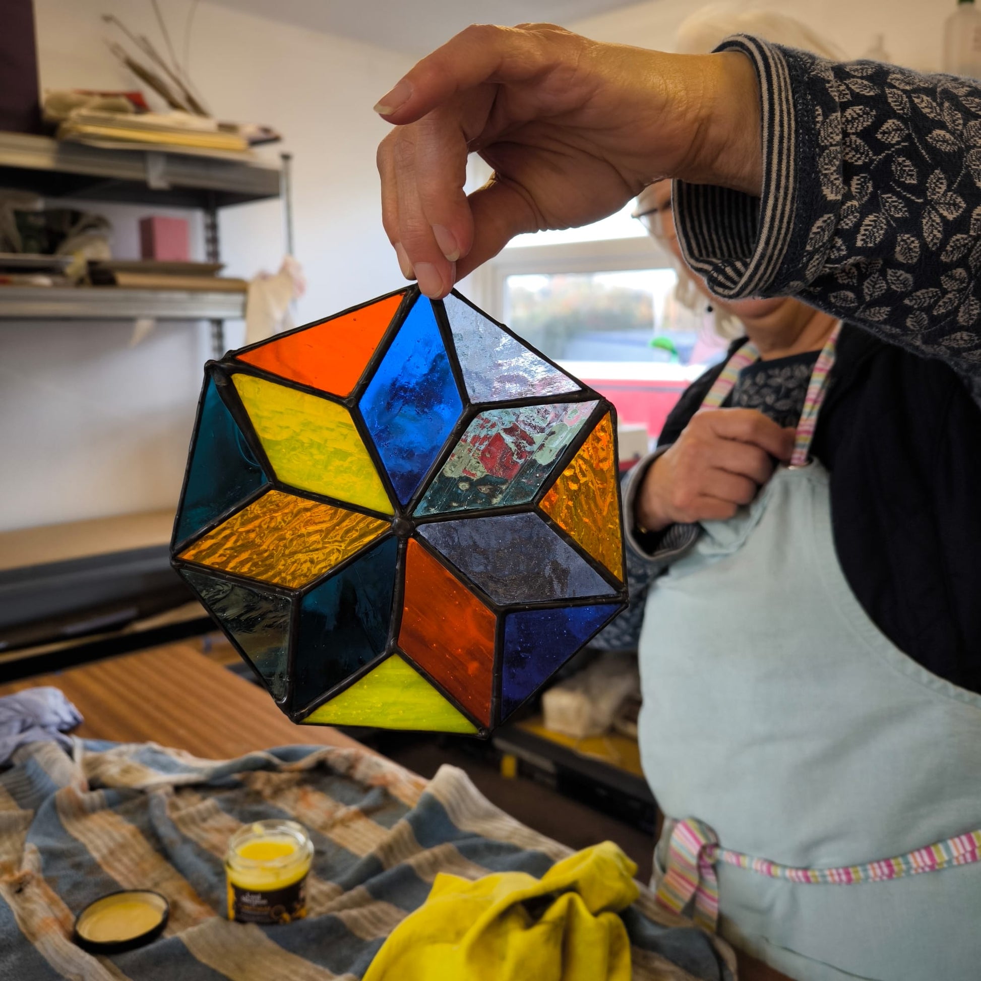 In a stained glass studio, an individual clad in a red hoodie and blue knit hat is focused on creating a vibrant stained glass panel featuring trees. The text reads, "10 Week Stained Glass Course (In Person), Tuesday EVENING by LF Kaleidoscope. Explore copper foiling, leaded stained glass and glass painting. 