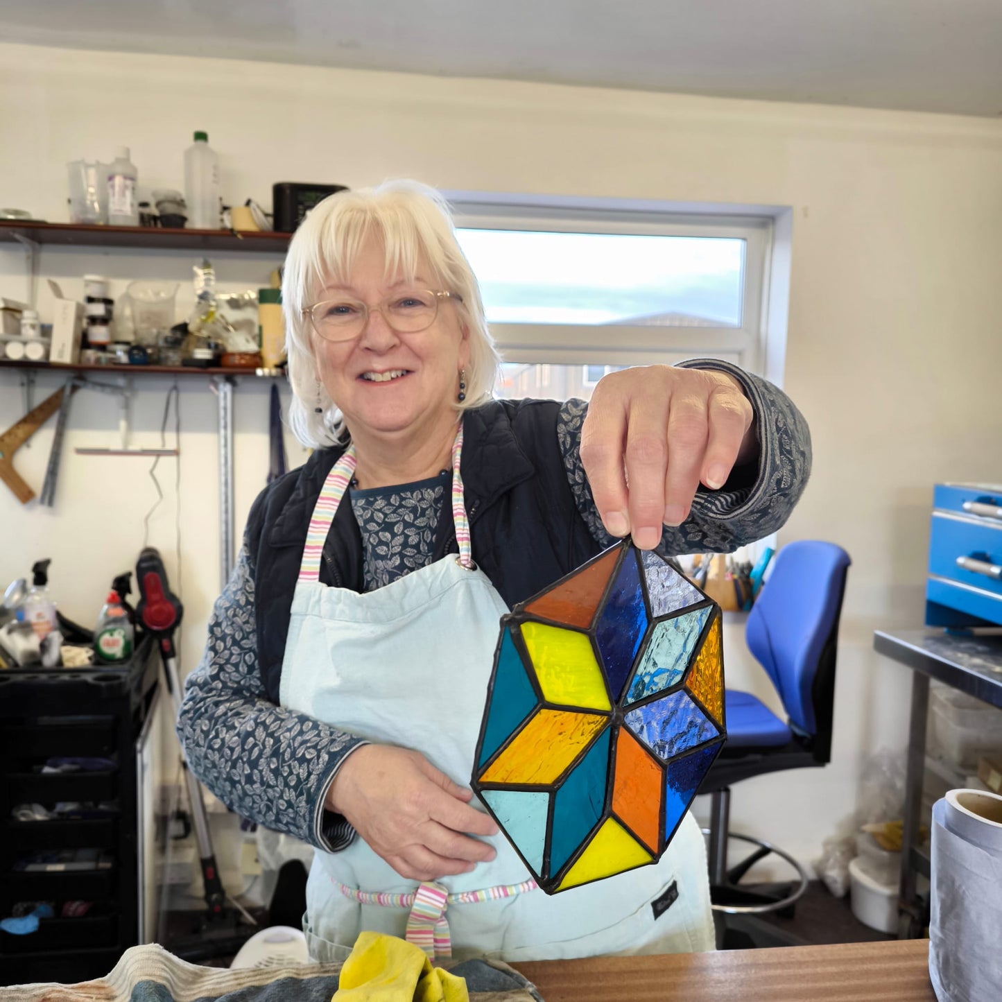 In a stained glass studio, an individual clad in a red hoodie and blue knit hat is focused on creating a vibrant stained glass panel featuring trees. The text reads, "10 Week Stained Glass Course (In Person), Tuesday EVENING by LF Kaleidoscope. Explore copper foiling, leaded stained glass and glass painting. 