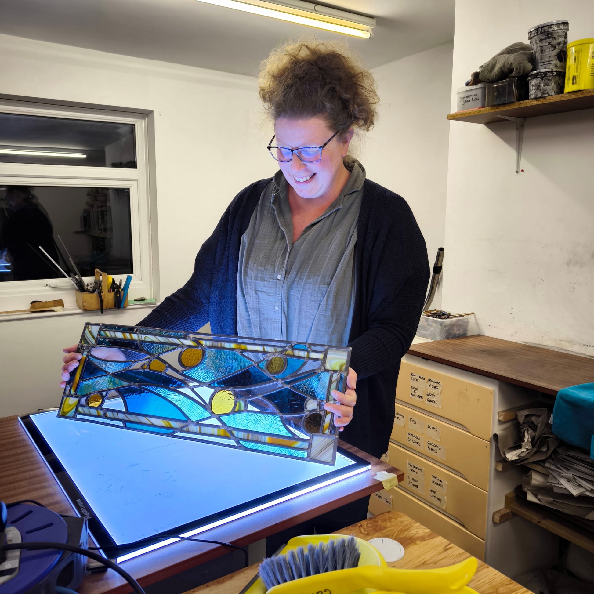 In a stained glass studio, an individual clad in a red hoodie and blue knit hat is focused on creating a vibrant stained glass panel featuring trees. The text reads, "10 Week Stained Glass Course (In Person), Tuesday EVENING by LF Kaleidoscope. Explore copper foiling, leaded stained glass and glass painting. 