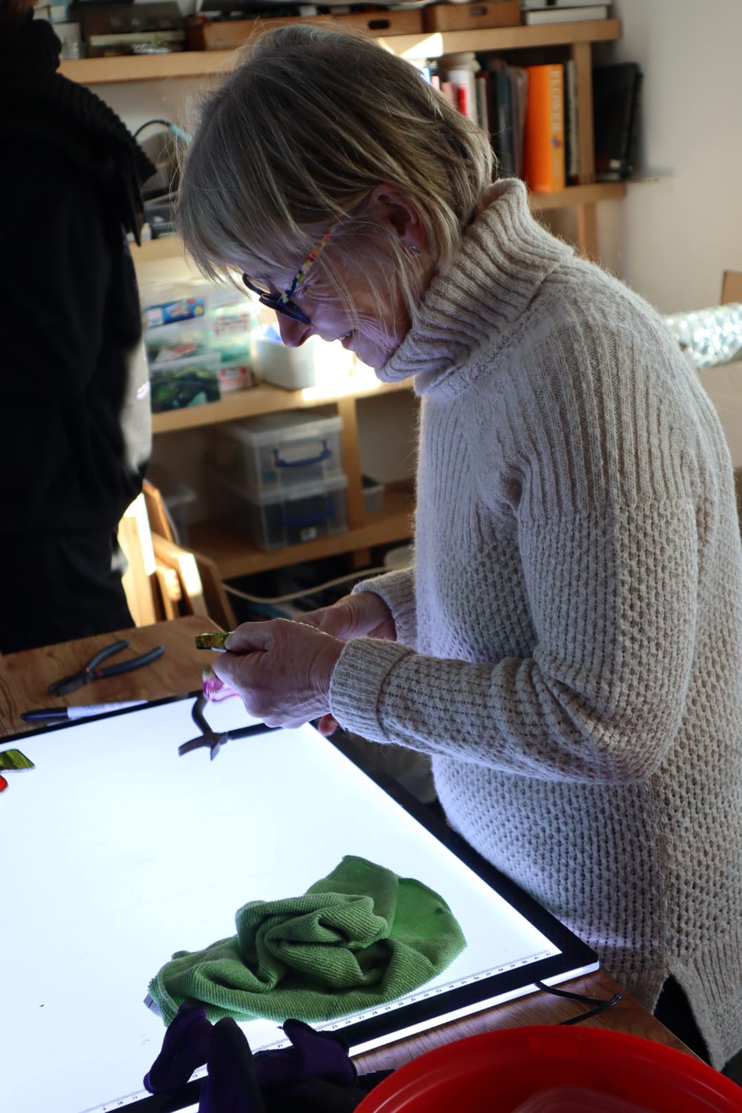 In a stained glass studio, an individual clad in a red hoodie and blue knit hat is focused on creating a vibrant stained glass panel featuring trees. The text reads, "10 Week Stained Glass Course (In Person), Tuesday EVENING by LF Kaleidoscope. Explore copper foiling, leaded stained glass and glass painting. 