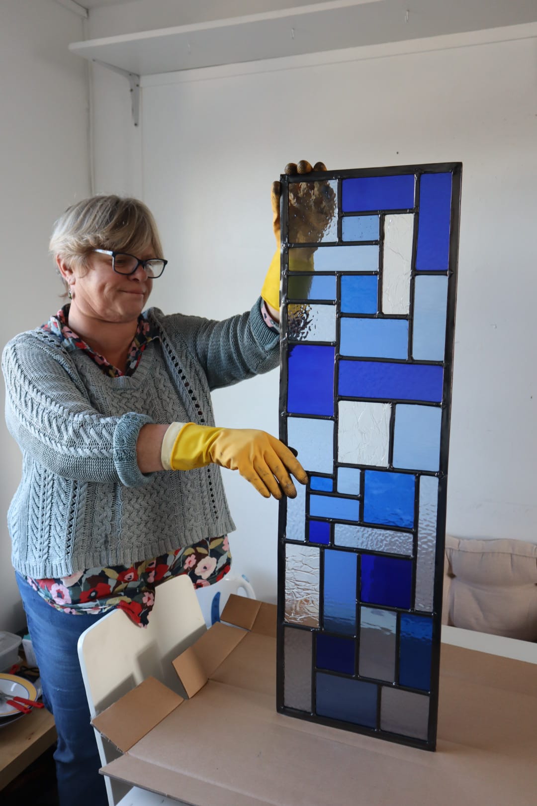 In a stained glass studio, an individual clad in a red hoodie and blue knit hat is focused on creating a vibrant stained glass panel featuring trees. The text reads, "10 Week Stained Glass Course (In Person), Tuesday EVENING by LF Kaleidoscope. Explore copper foiling, leaded stained glass and glass painting. 