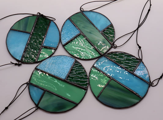 Four distinct pieces from the "Circles, Green" collection by LF Kaleidoscope are showcased against a white backdrop. Each suspended circle displays geometric patterns in hues of green and blue, with diverse textures. They are affixed to black strings for hanging.