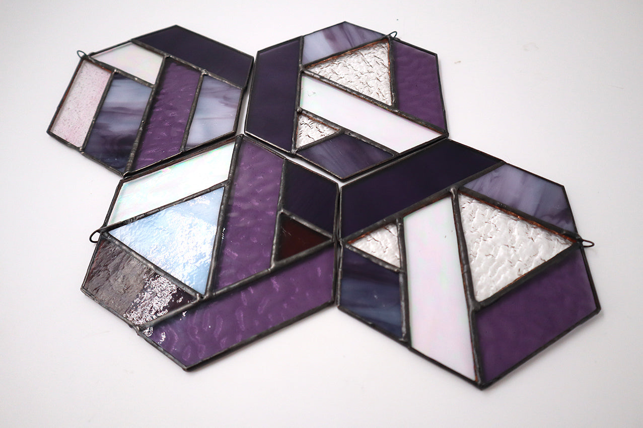 Introducing the "Hexagons, Purple" by LF Kaleidoscope: a stunning geometric-inspired stained glass ornament crafted from vintage glass offcuts. This hexagonal piece showcases a mix of textured lavender, iridescent, and solid shades of purple that hang beautifully against any plain background.