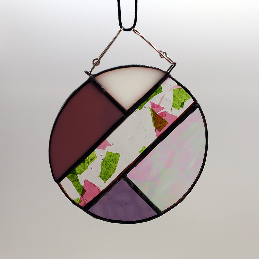 This unique creation from LF Kaleidoscope, known as "Circles, Purple," is a circular stained glass piece showcasing sections of purple, white, and brown glass skillfully arranged with intersecting green geometric patterns. Delicate hanging circles are suspended from hooks against a plain white backdrop, offering a stunning display of artistry.