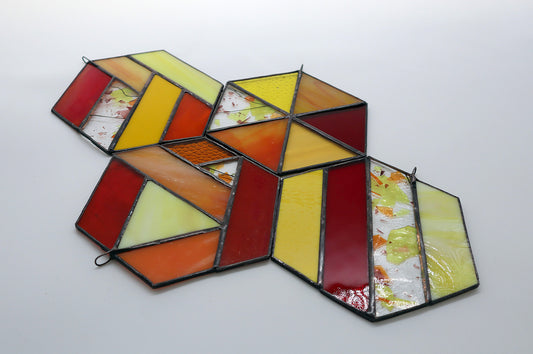 A geometric stained glass piece composed of hexagonal sections featuring red, yellow, orange, and clear glass segments in various patterns and textures, laid on a white background.