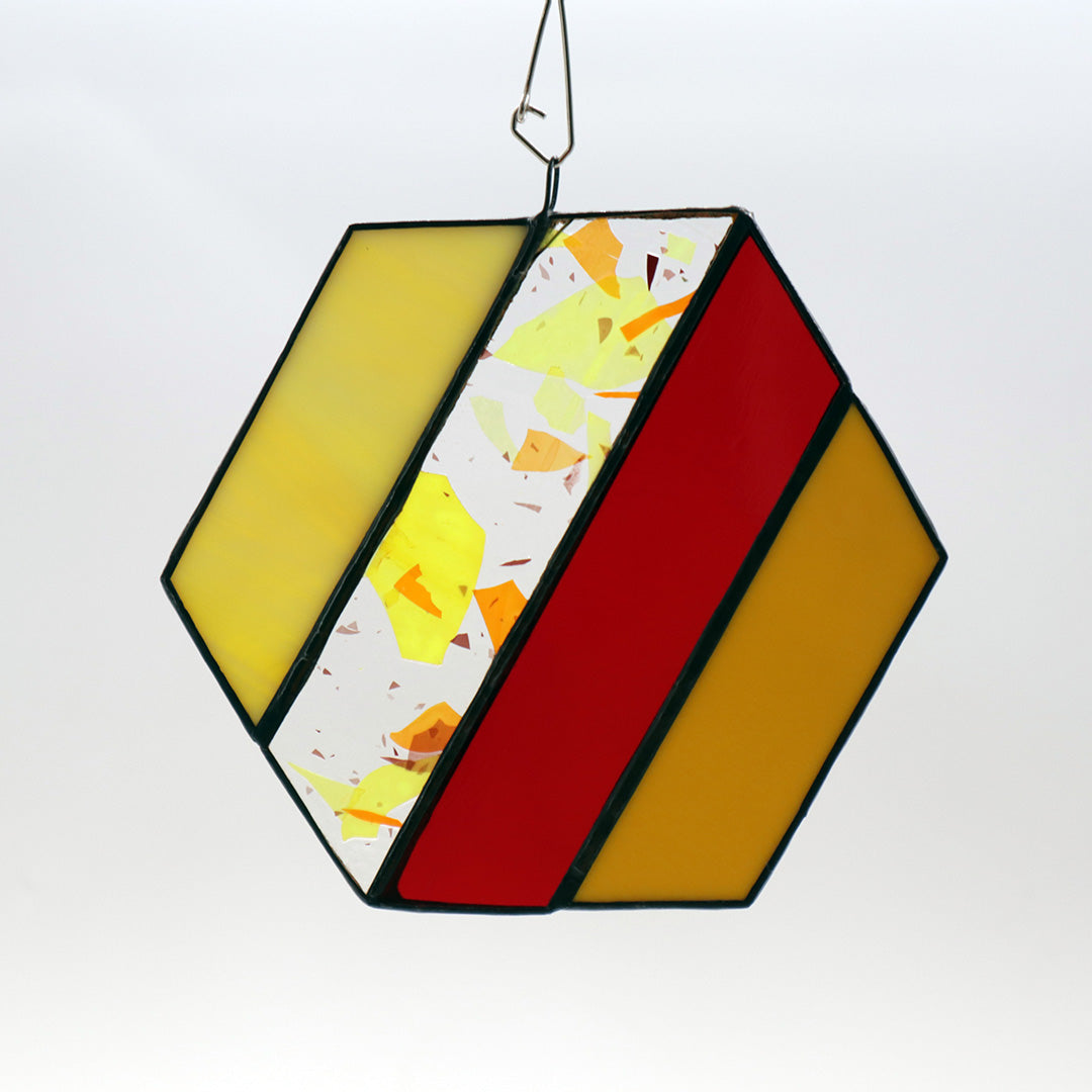 Introducing "Hexagons, Orange" by LF Kaleidoscope—a vintage glass hexagonal stained ornament showcasing a geometric design with vibrant sections of yellow, red, and white, enhanced with delicate orange flecks. Framed in a dark border and elegantly suspended by a wire, this homemade decor piece brings artistic flair to any space.