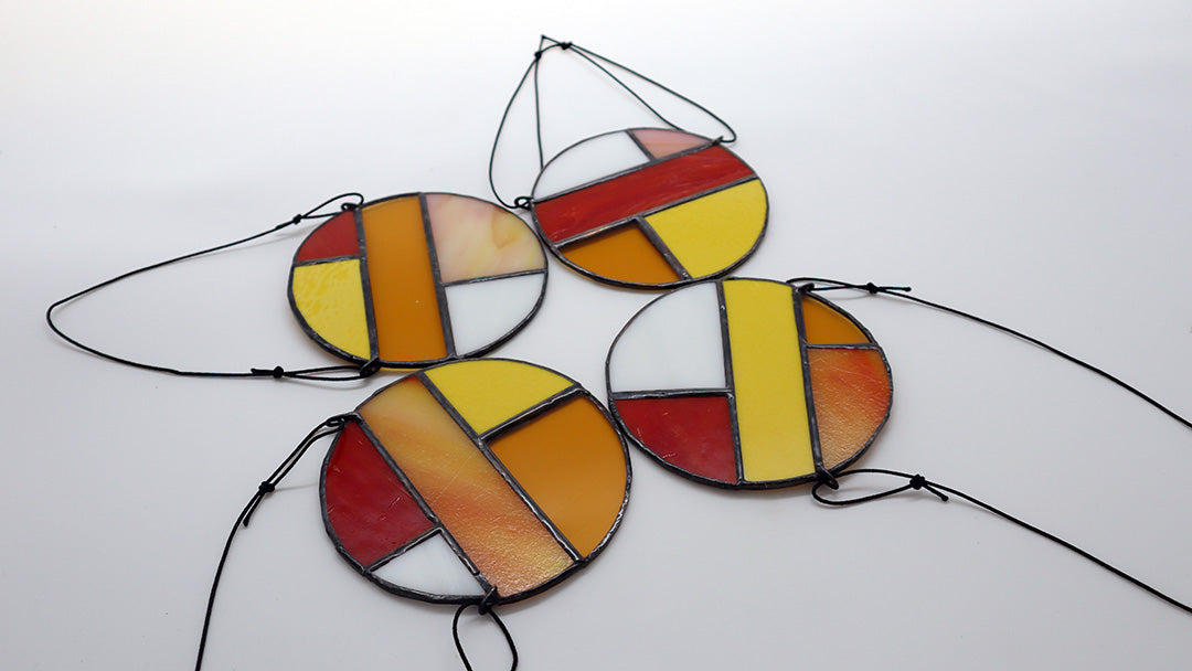 Featuring four distinct pieces, these stained glass circles by LF Kaleidoscope exhibit geometric designs in shades of red, yellow, orange, and white. Each circle is elegantly framed in black and equipped with strings for effortless hanging. They are artistically displayed on a light surface to enhance their vibrant colors.