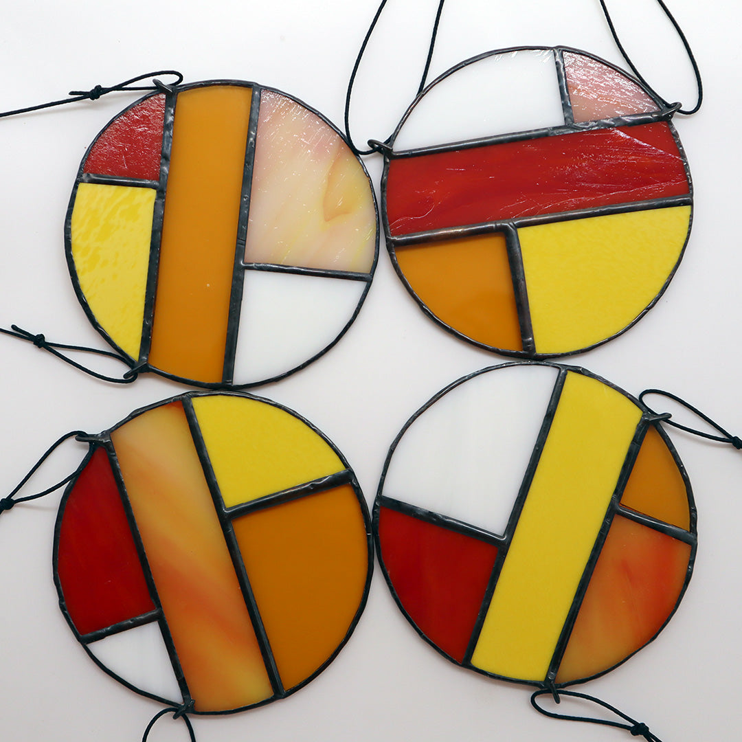 Featuring four distinct pieces, these stained glass circles by LF Kaleidoscope exhibit geometric designs in shades of red, yellow, orange, and white. Each circle is elegantly framed in black and equipped with strings for effortless hanging. They are artistically displayed on a light surface to enhance their vibrant colors.