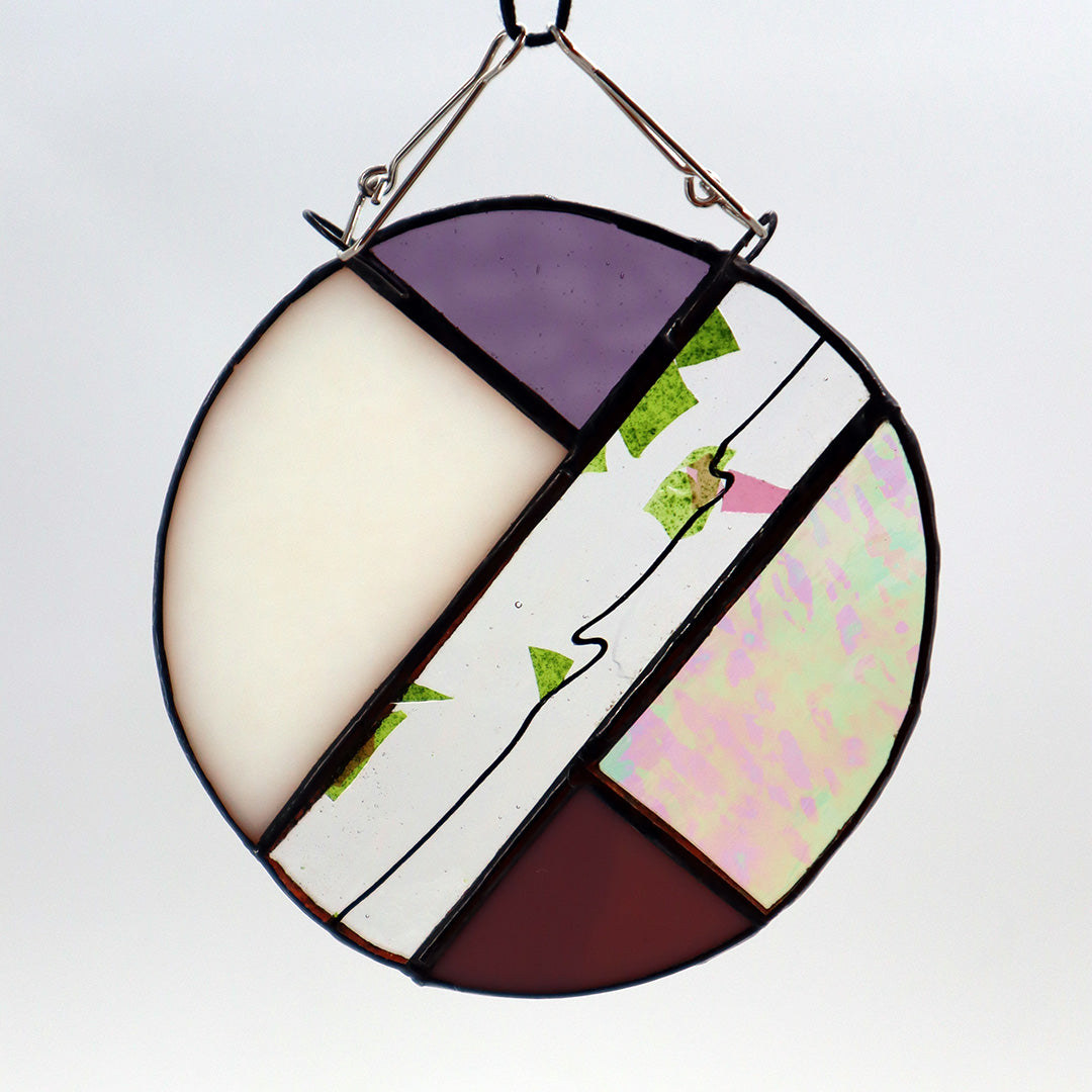 This unique creation from LF Kaleidoscope, known as "Circles, Purple," is a circular stained glass piece showcasing sections of purple, white, and brown glass skillfully arranged with intersecting green geometric patterns. Delicate hanging circles are suspended from hooks against a plain white backdrop, offering a stunning display of artistry.