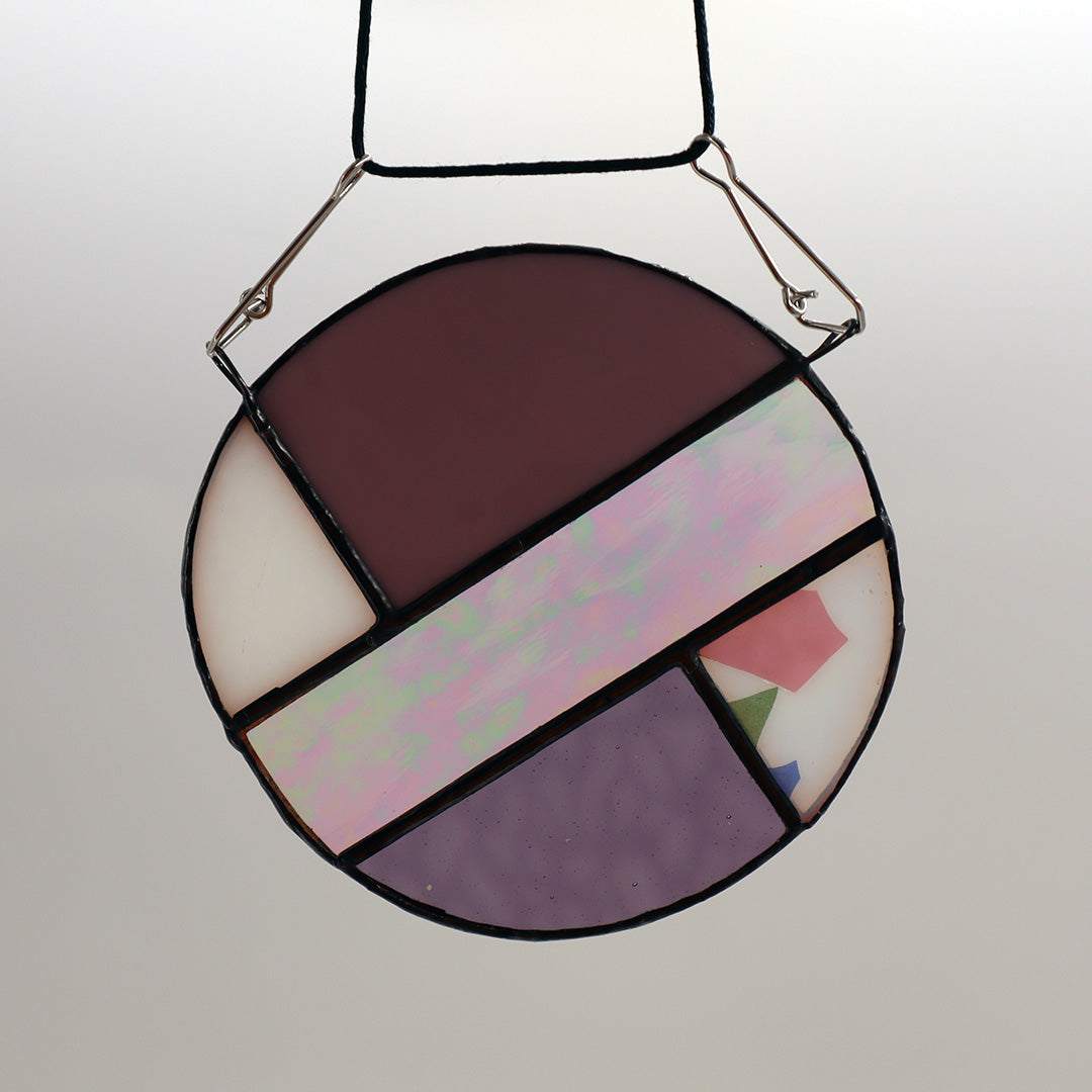 This unique creation from LF Kaleidoscope, known as "Circles, Purple," is a circular stained glass piece showcasing sections of purple, white, and brown glass skillfully arranged with intersecting green geometric patterns. Delicate hanging circles are suspended from hooks against a plain white backdrop, offering a stunning display of artistry.