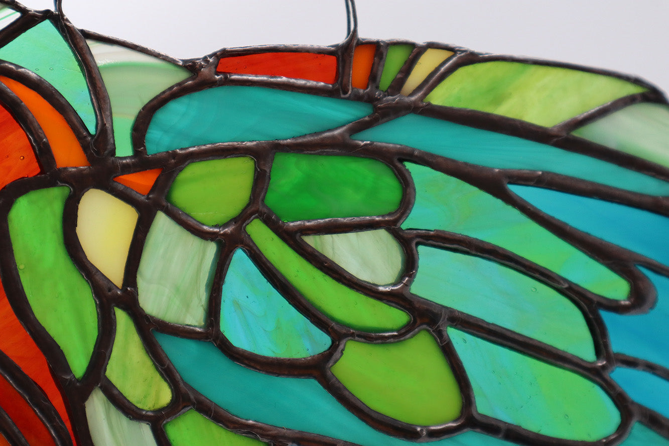 Close up of the wing and feathers of: 
The "Mr Feathers" handmade glass art piece by LF Kaleidoscope beautifully presents a stained glass depiction of a parrot. The vibrant colours of green, orange, blue, and turquoise are intricately outlined in black copperfoil work to define the bird's striking details, making it perfect as a decorative window hanging against a light neutral background. The face and eyes are exquisitely painted and really bring this stunning collector piece to life. 