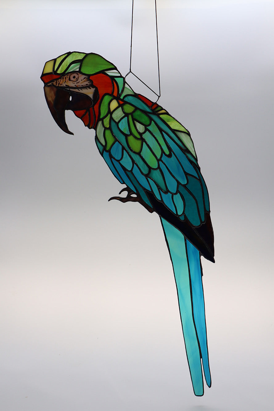 The "Mr Feathers" handmade glass art piece by LF Kaleidoscope beautifully presents a stained glass depiction of a parrot. The vibrant colours of green, orange, blue, and turquoise are intricately outlined in black copperfoil work to define the bird's striking details, making it perfect as a decorative window hanging against a light neutral background. The face and eyes are exquisitely painted and really bring this stunning collector piece to life. 