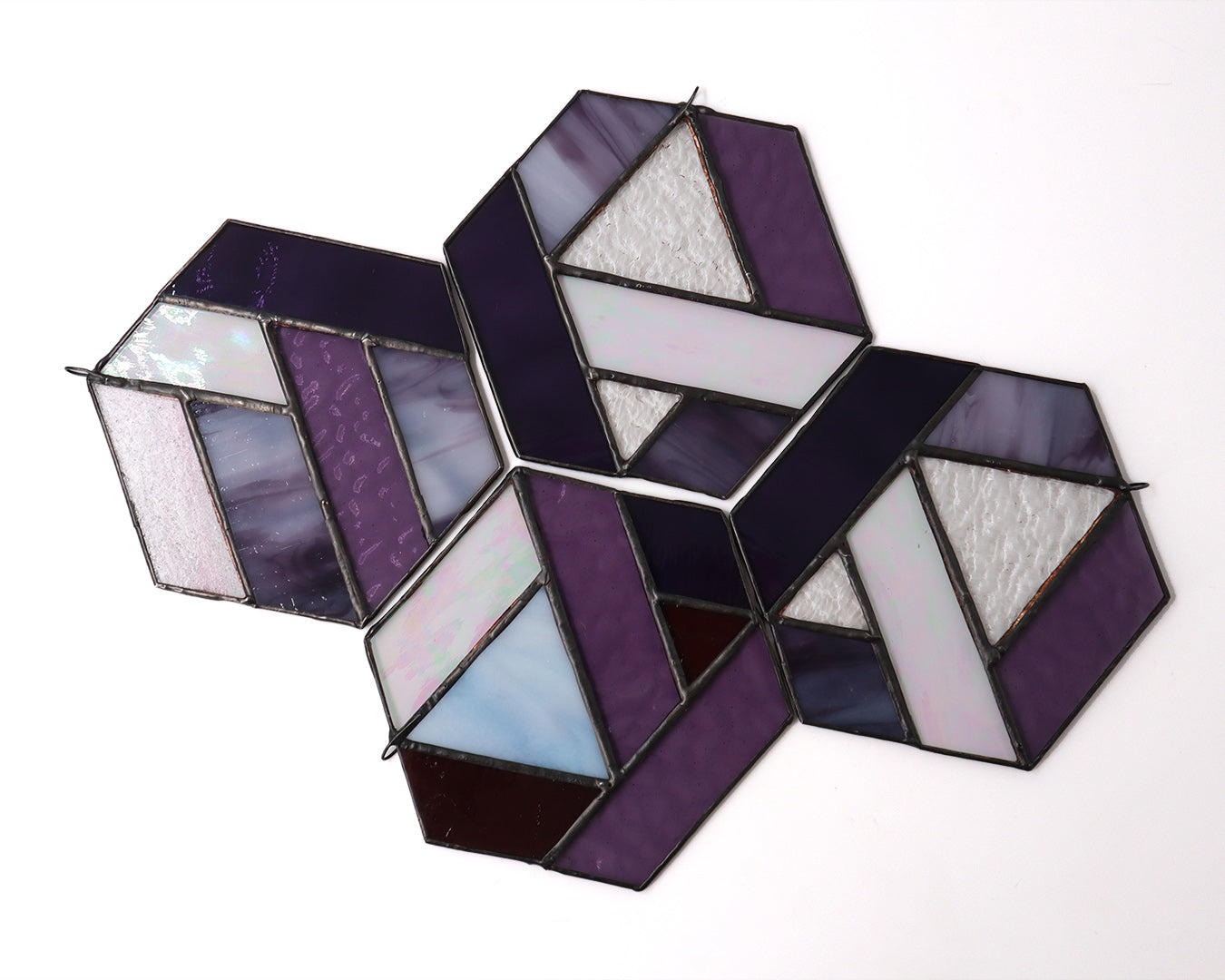 Introducing the "Hexagons, Purple" by LF Kaleidoscope: a stunning geometric-inspired stained glass ornament crafted from vintage glass offcuts. This hexagonal piece showcases a mix of textured lavender, iridescent, and solid shades of purple that hang beautifully against any plain background.