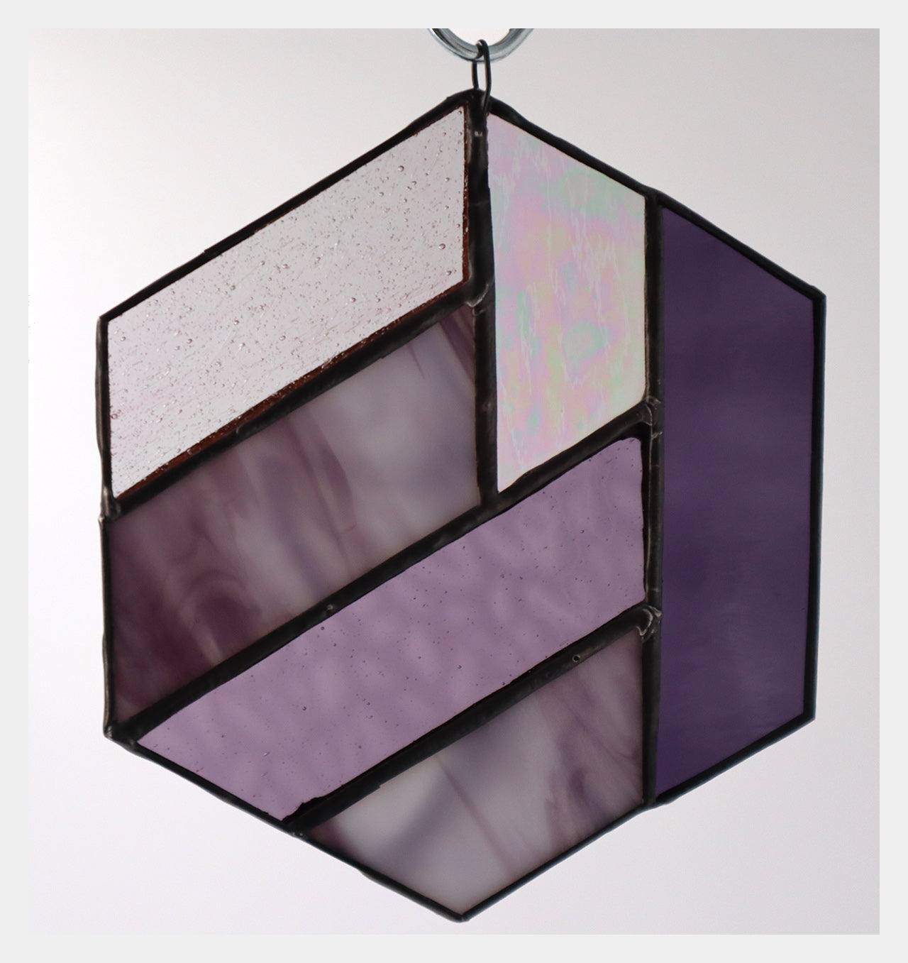 Introducing the "Hexagons, Purple" by LF Kaleidoscope: a stunning geometric-inspired stained glass ornament crafted from vintage glass offcuts. This hexagonal piece showcases a mix of textured lavender, iridescent, and solid shades of purple that hang beautifully against any plain background.