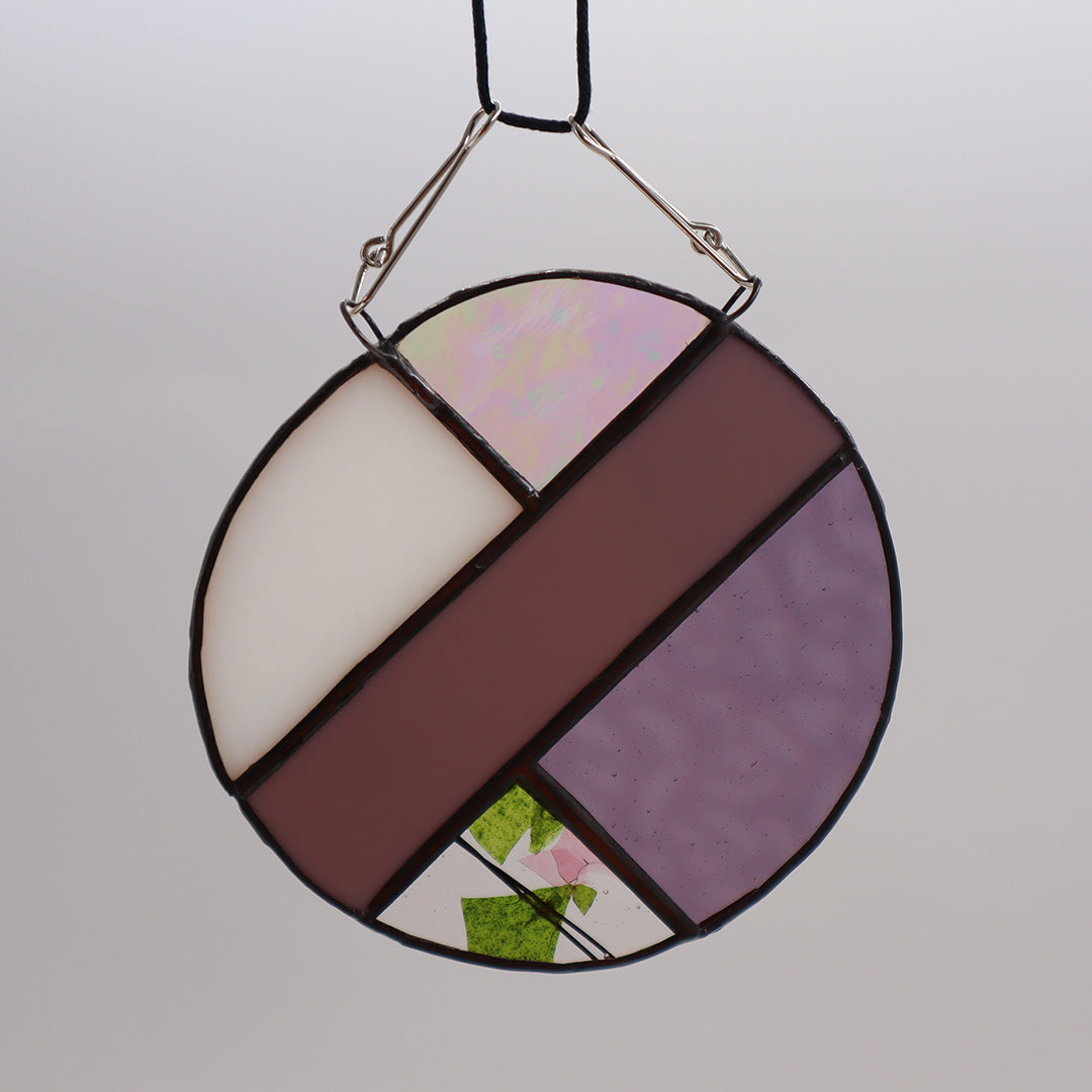 This unique creation from LF Kaleidoscope, known as "Circles, Purple," is a circular stained glass piece showcasing sections of purple, white, and brown glass skillfully arranged with intersecting green geometric patterns. Delicate hanging circles are suspended from hooks against a plain white backdrop, offering a stunning display of artistry.