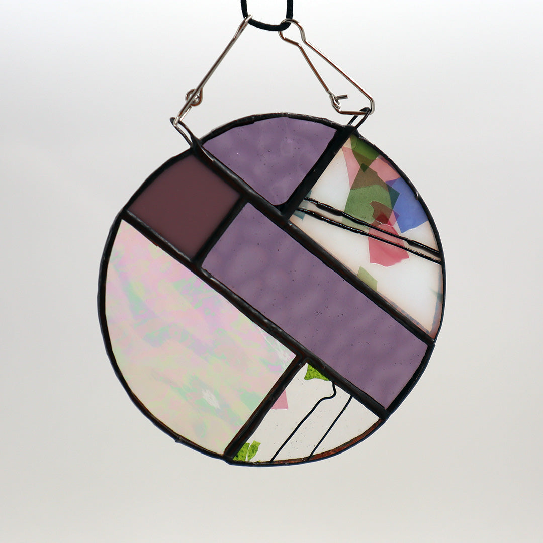 This unique creation from LF Kaleidoscope, known as "Circles, Purple," is a circular stained glass piece showcasing sections of purple, white, and brown glass skillfully arranged with intersecting green geometric patterns. Delicate hanging circles are suspended from hooks against a plain white backdrop, offering a stunning display of artistry.