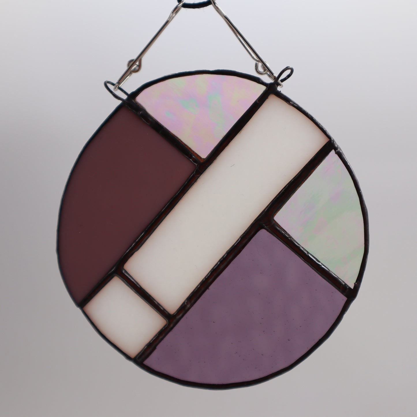 This unique creation from LF Kaleidoscope, known as "Circles, Purple," is a circular stained glass piece showcasing sections of purple, white, and brown glass skillfully arranged with intersecting green geometric patterns. Delicate hanging circles are suspended from hooks against a plain white backdrop, offering a stunning display of artistry.